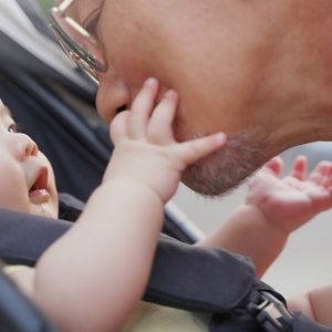 Baby in car seat looks at father and touches father's face.