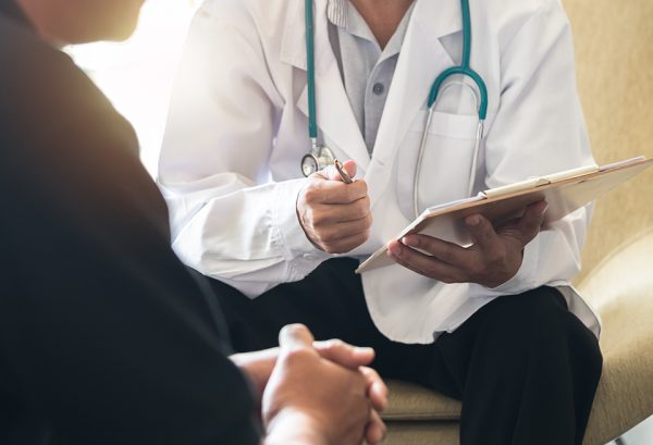 A doctor holding a clipboard and pen talks to a male patient.