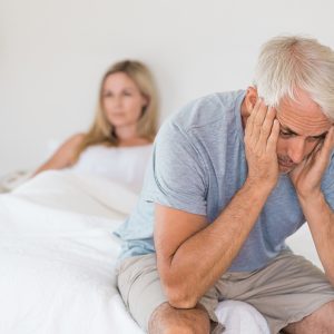 A man sits on the edge of a bed with his hands cradling his face. A woman sits under the covers in the background.