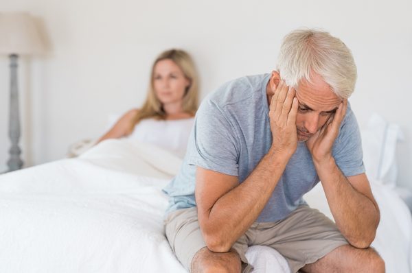 A man sits on the edge of a bed with his hands cradling his face. A woman sits under the covers in the background.